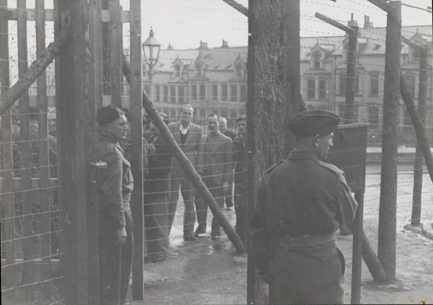 Schwarzweißfotografie. Uniformierte vor Stacheldrahtzaun, dahinter mehrere zivil gekleidete Männer.