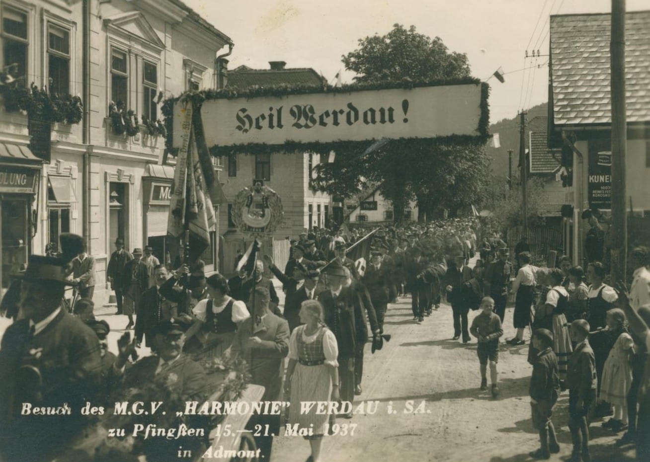 Historic recording of a festival procession. Several people are carrying flags with swastikas. A banner hangs above them with the inscription "Heil Werdau!".